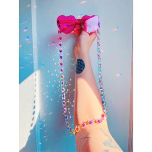 May include: Pink heart-shaped sunglasses with a matching beaded chain. The sunglasses are held up, showcasing the iridescent chain and the reflection of light. The chain is made of colorful, iridescent beads.