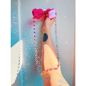 May include: Heart-shaped sunglasses with bright pink lenses and a silver-toned metal chain strap featuring iridescent, multi-colored faceted beads. The chain hangs down from the frames, creating a long, decorative accessory for eyewear.