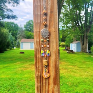 Può includere: Un ornamento da appendere con una catenella color argento, tre perline in legno testurizzato, un ciondolo circolare in metallo con motivo floreale geometrico, due cristalli iridati a goccia, piccole perline arancioni e verdi e una fetta di agata arancione.