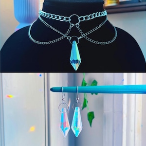 May include: A silver-toned choker necklace with a layered design and a dangling iridescent crystal pendant. Below, a pair of matching earrings with similar crystal pendants hang from silver hooks. The jewelry is displayed on a black mannequin head and a blue rod.