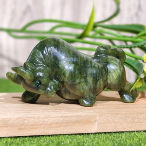 May include: A green jade bull figurine, sculpted in a charging pose. The bull has detailed features and is set on a wooden surface, with a sunflower and greenery in the background. The figurine is approximately 5 inches long.