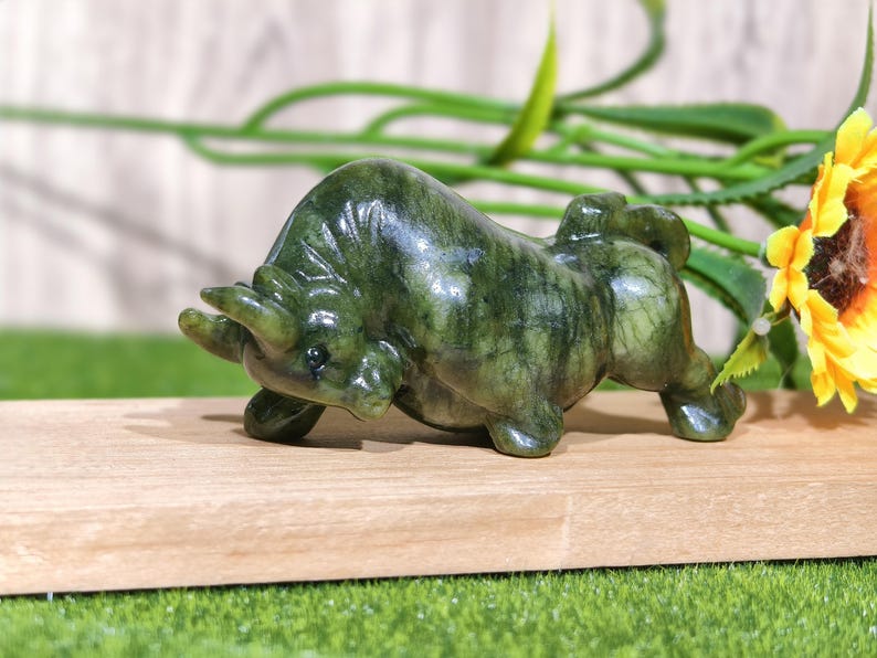 May include: A green jade bull figurine, sculpted in a charging pose. The bull has detailed features and is set on a wooden surface, with a sunflower and greenery in the background. The figurine is approximately 5 inches long.