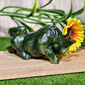 May include: A dark green jade bull figurine with a textured surface. The bull is lying down on a wooden surface, with a yellow sunflower and green foliage in the background.