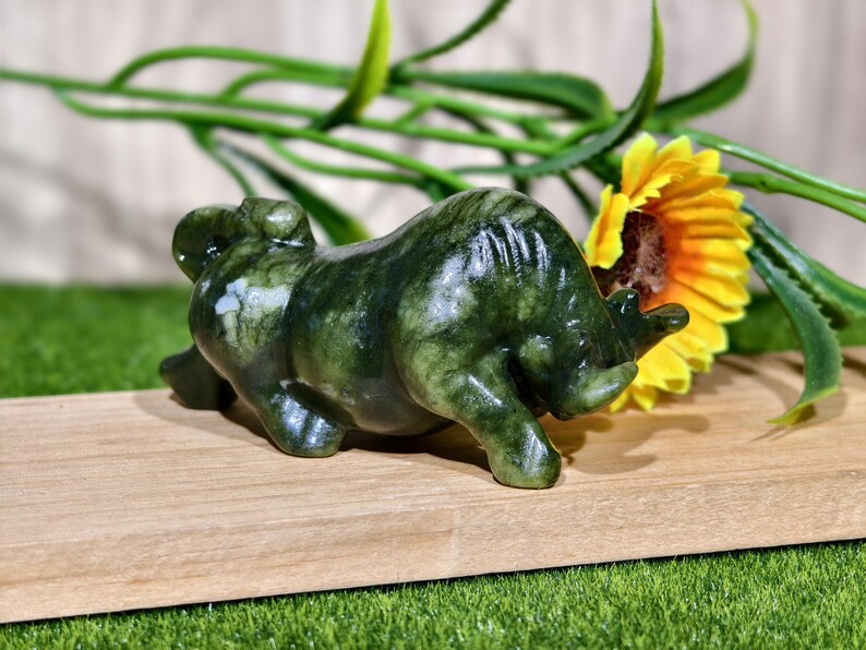 May include: A dark green jade bull figurine with a textured surface. The bull is lying down on a wooden surface, with a yellow sunflower and green foliage in the background.