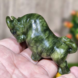 May include: A small, green stone sculpture of a bull. The bull is carved with detailed features and has a polished, glossy finish. The sculpture is held in a person's hand, showcasing its size and form. The stone has a natural, marbled appearance.