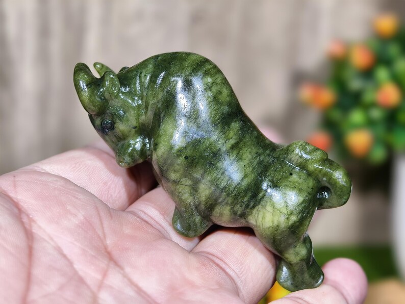 May include: A small, green stone sculpture of a bull. The bull is carved with detailed features and has a polished, glossy finish. The sculpture is held in a person's hand, showcasing its size and form. The stone has a natural, marbled appearance.