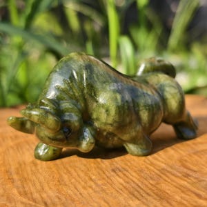 May include: A green and brown stone bull figurine, sculpted in a realistic pose. The bull is on all fours, with its head lowered and horns visible. The figurine is set on a wooden surface.