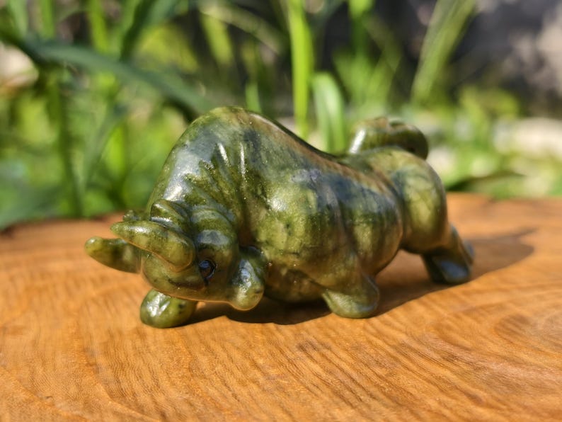 May include: A green and brown stone bull figurine, sculpted in a realistic pose. The bull is on all fours, with its head lowered and horns visible. The figurine is set on a wooden surface.