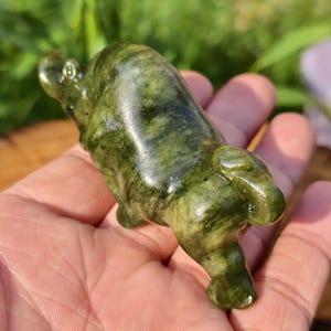 May include: A carved green stone figurine of a hippo. The hippo is lying down with its legs tucked under its body. The stone has a mottled pattern of dark and light green.