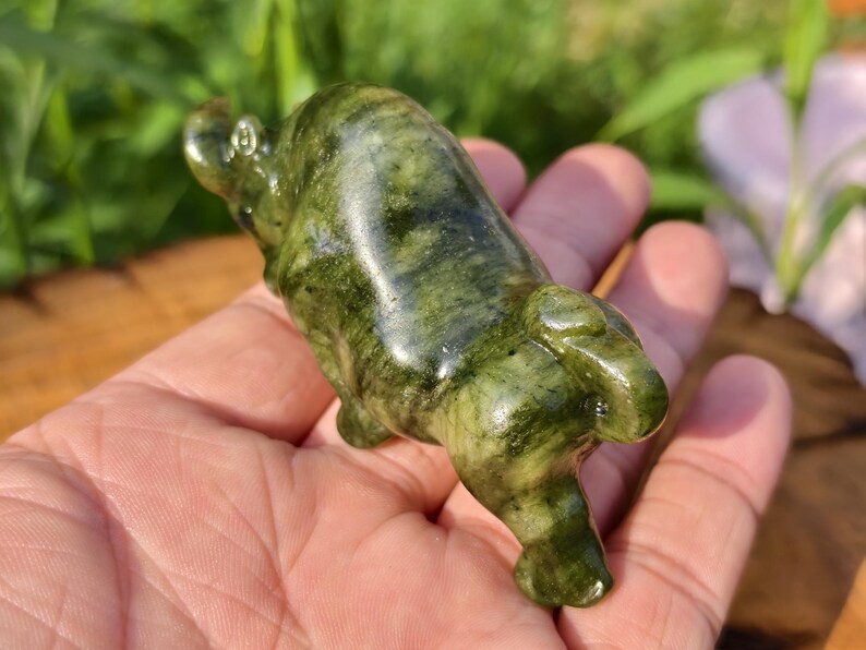 May include: A carved green stone figurine of a hippo. The hippo is lying down with its legs tucked under its body. The stone has a mottled pattern of dark and light green.