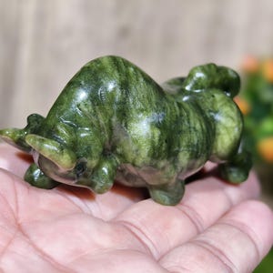 May include: A small, green stone sculpture of a bull. The bull is carved with detailed features, including horns and muscular body. The stone has a marbled appearance with varying shades of green.