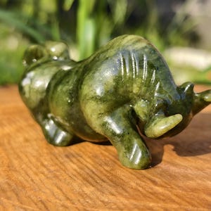May include: A green jade bull figurine with a polished finish. The sculpture features detailed carving and a natural color variation. The bull is posed in a charging stance, showcasing its horns and muscular build. A decorative item.