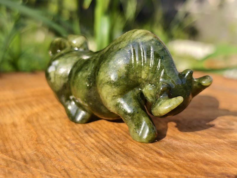 May include: A green jade bull figurine with a polished finish. The sculpture features detailed carving and a natural color variation. The bull is posed in a charging stance, showcasing its horns and muscular build. A decorative item.