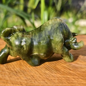 May include: A jade figurine of a bull in a charging pose. The sculpture is a mottled green color with white veining, and is set on a wooden surface. The bull has detailed features and a polished finish.