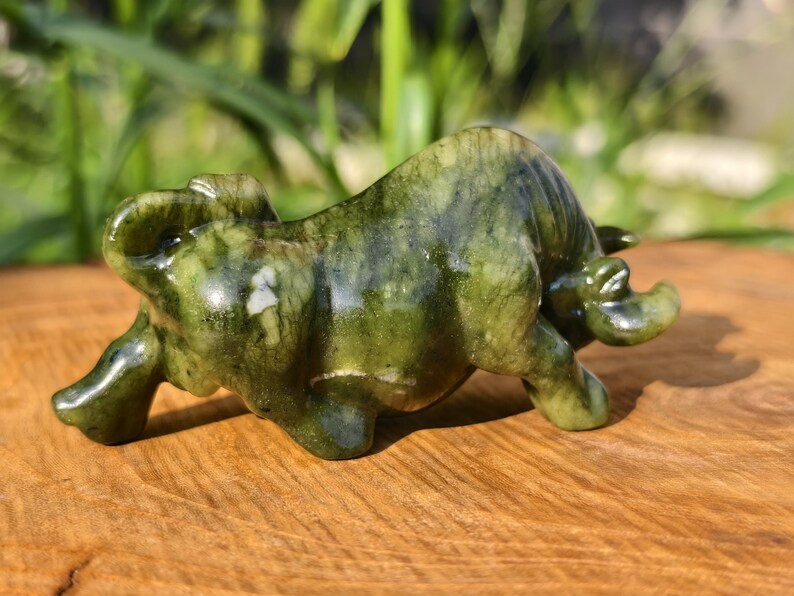 May include: A jade figurine of a bull in a charging pose. The sculpture is a mottled green color with white veining, and is set on a wooden surface. The bull has detailed features and a polished finish.