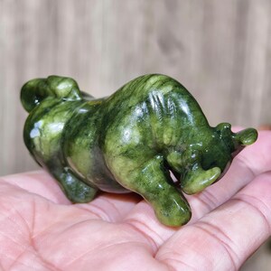 May include: A green jade bull figurine. The sculpture is a detailed representation of a bull, with a polished, glossy finish. The jade has varying shades of green, creating a natural, marbled effect. The bull is posed in a charging stance.