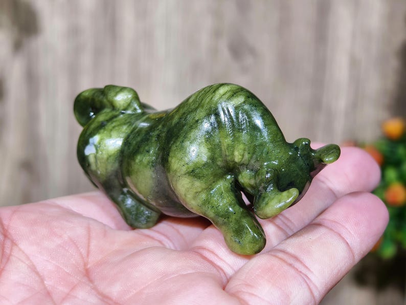 May include: A green jade bull figurine. The sculpture is a detailed representation of a bull, with a polished, glossy finish. The jade has varying shades of green, creating a natural, marbled effect. The bull is posed in a charging stance.
