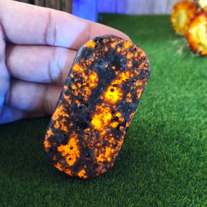 Puede incluir: Una piedra rectangular pulida con un llamativo patrón de motas negras y naranja vibrante. La superficie de la piedra es lisa y los colores crean un efecto ardiente y brillante. La piedra se sostiene sobre un fondo verde.