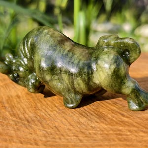May include: A green and brown carved stone bull figurine. The sculpture is lying down on a wooden surface, with its head lowered and legs extended. The bull has a glossy finish and is set against a blurred green background.