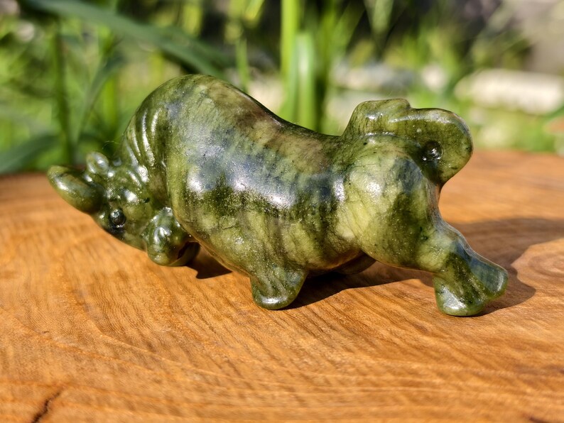 May include: A green and brown carved stone bull figurine. The sculpture is lying down on a wooden surface, with its head lowered and legs extended. The bull has a glossy finish and is set against a blurred green background.