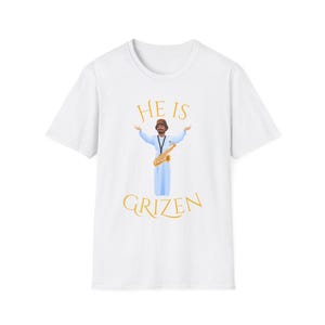 Könnte beinhalten: Weißes T-Shirt mit einer Grafik einer Figur, die Saxophon spielt, mit dem Text "HE IS GRIZEN" in Gold. Die Figur trägt eine blaue Robe und hat die Arme ausgestreckt. Das T-Shirt ist aus einem weichen, bequemen Material.