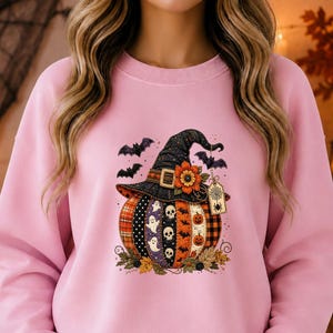 Peut inclure: Un sweat-shirt à col rond rose clair orné d'un motif central représentant une citrouille portant un chapeau de sorcière. La citrouille est décorée de bandes verticales avec des crânes, des fantômes et des motifs à carreaux. Des chauves-souris volent autour du chapeau.