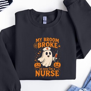 Może przedstawiać: Czarna bluza z grafiką o tematyce Halloween. Projekt przedstawia ducha w czepku pielęgniarki i strzykawką, z napisem "MY BROOM BROKE so now I'm a NURSE". Dwie pomarańczowe dynie po obu stronach tekstu. Zabawny, sezonowy element garderoby.