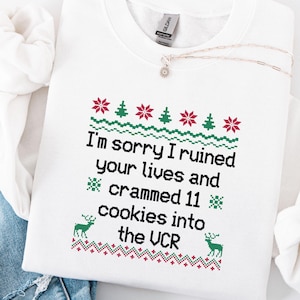 Może przedstawiać: Biała bluza z okrągłym dekoltem z świątecznym wzorem. Wzór zawiera pikselowy wzór czerwonych i zielonych choinek, płatków śniegu i reniferów. Tekst brzmi: „Przepraszam, że zrujnowałem wam życie i wepchnąłem 11 ciasteczek do magnetowidu”.