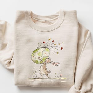 May include: A beige sweatshirt featuring a watercolor illustration of a rabbit carrying a large, cracked egg adorned with flowers. The egg has a green and white design. The rabbit is brown and white, standing on a grassy area.