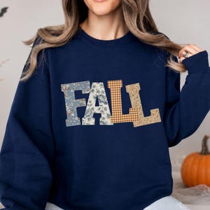 May include: A navy blue crewneck sweatshirt featuring the word FALL in large block letters across the chest. The letters F and A have a blue floral pattern, while the letters L and L feature a brown and white gingham and gold glitter texture.