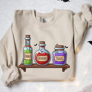 Może przedstawiać: Beżowa bluza z motywem Halloween. Projekt zawiera trzy butelki z miksturami z etykietami "Fangs", "Demons" i "Toads" na drewnianej półce. Czarne nietoperze są rozrzucone.