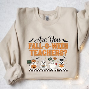 May include: Beige sweatshirt with the text "Are You Fall-O-Ween Teachers?" and illustrations of ghosts dressed as teachers. The ghosts are holding books, apples, and a chalkboard. Black bats are scattered around the top.