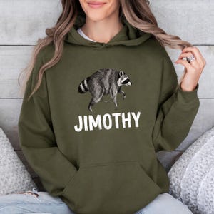 May include: An olive green hooded sweatshirt featuring a graphic illustration of a gray and black raccoon in the center. Below the raccoon, the text JIMOTHY is printed in bold, white, capital letters. The hoodie includes a front kangaroo pocket.