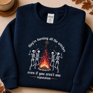 May include: A navy blue crewneck sweatshirt featuring a graphic of four skeletons dancing around a campfire. The text reads, "they're burning all the witches even if you aren't one reputation," with a small star and crescent moon symbol above the fire.