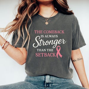 Könnte beinhalten: Dunkelgraues T-Shirt mit dem Text "THE COMEBACK IS ALWAYS STRONGER THAN THE SETBACK" in Weiß und Rosa, sowie einer rosa Schleife. Das T-Shirt hat einen lässigen Stil.