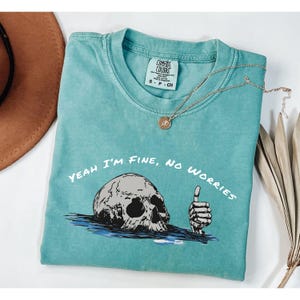Può includere: T-shirt turchese Comfort Colors con la scritta "Yeah I'm Fine, No Worries" e una grafica di un teschio e una mano con il pollice in su. Sono visibili anche una collana d'oro e un cappello marrone.