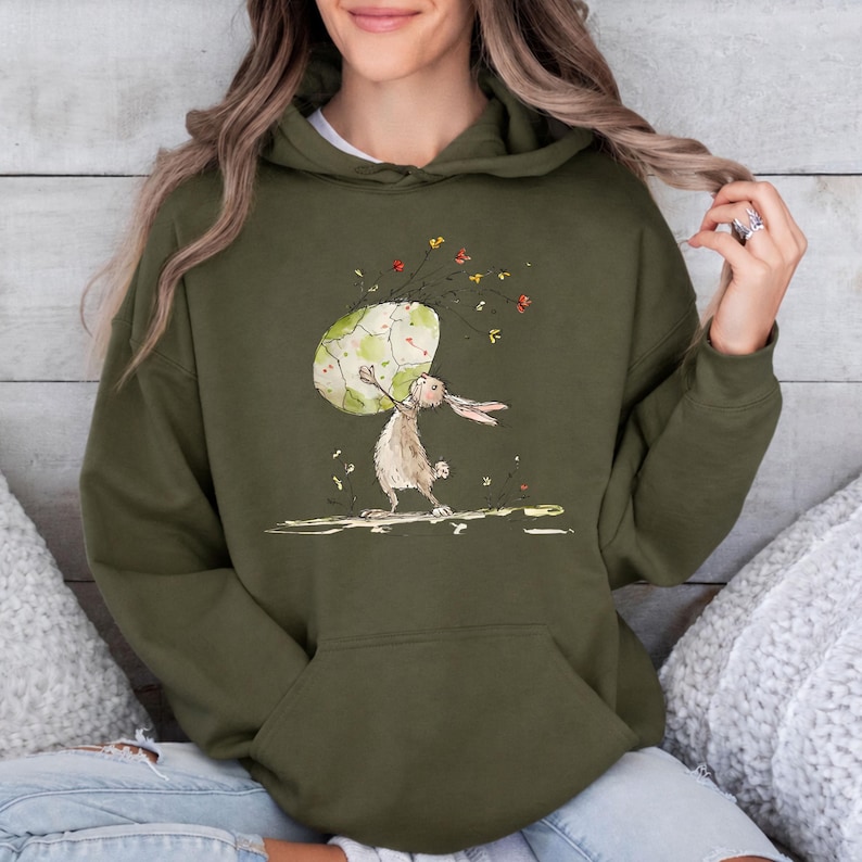 May include: An olive green hooded sweatshirt featuring a graphic illustration of a brown rabbit holding a large, speckled, light green egg. The design includes small, colorful floral accents floating above the egg and a simple ground line.