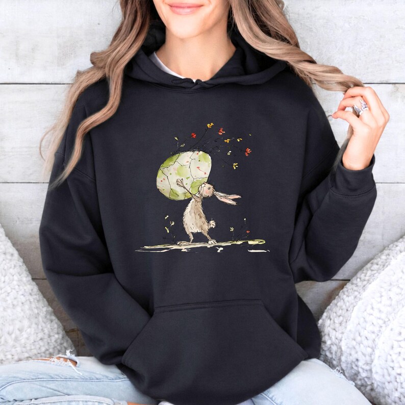 May include: A black hooded sweatshirt featuring a graphic illustration of a brown rabbit standing on its hind legs while holding a large, textured, light green egg. Small, colorful leaf-like shapes are scattered around the egg and rabbit.