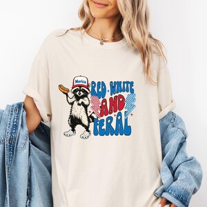 K&ouml;nnte beinhalten: Cremefarbenes T-Shirt mit einem Waschb&auml;ren, der einen Hut mit der Aufschrift "Merica" tr&auml;gt und einen Hotdog h&auml;lt. Das Shirt hat den Text "Red, White, and Feral" in Rot, Wei&szlig; und Blau mit Blitzgrafiken. Das Shirt ist ein l&auml;ssiger Stil.