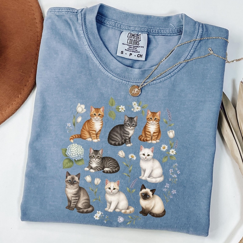 May include: A light blue Comfort Colors t-shirt featuring a design of various cat breeds surrounded by flowers. The cats are in different poses and colors, including orange, gray, and white. A gold necklace is draped over the shirt.