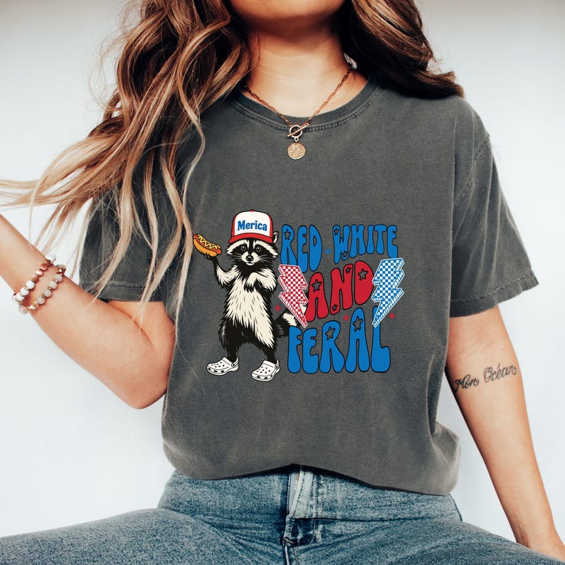 K&ouml;nnte beinhalten: Dunkelgraues T-Shirt mit einem Waschb&auml;ren, der einen Hut mit der Aufschrift "Merica" tr&auml;gt und einen Hotdog h&auml;lt. Das Shirt hat die Worte "Red White and Feral" in Rot und Blau mit Blitzgrafiken.