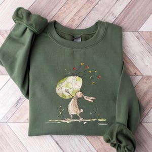 May include: An olive green crewneck sweatshirt featuring a watercolor-style illustration of a brown rabbit standing on its hind legs while holding a large, pale green and white patterned egg above its head, surrounded by small colorful wildflowers.