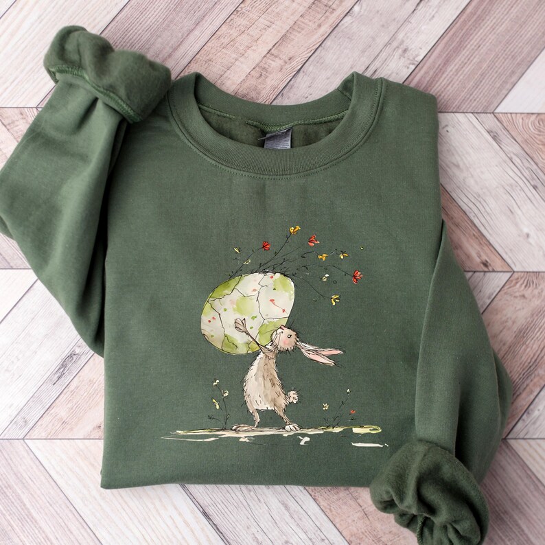 May include: An olive green crewneck sweatshirt featuring a watercolor-style illustration of a brown rabbit standing on its hind legs while holding a large, pale green and white patterned egg above its head, surrounded by small colorful wildflowers.