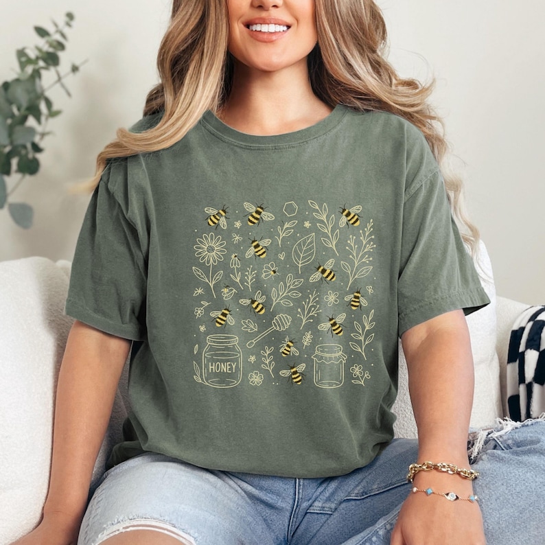May include: Olive green t-shirt featuring a square design with bees, flowers, leaves, and honey jars. The bees are yellow and black. The word "HONEY" is printed on one of the jars. The shirt is a short-sleeved, crew neck style.