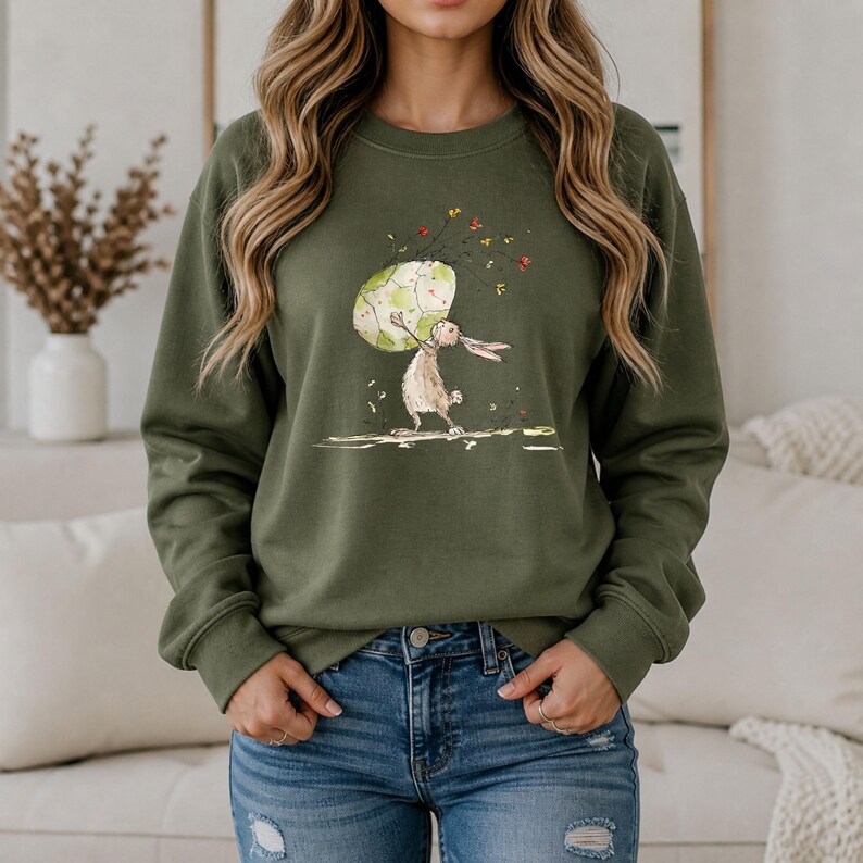 May include: An olive green crewneck sweatshirt featuring a graphic illustration of a light brown rabbit standing on its hind legs while holding a large, speckled, pale green egg above its head, surrounded by small, scattered red and yellow floral accents.