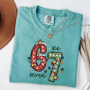 Puede incluir: Una camiseta turquesa Comfort Colors con los números "67" en un diseño navideño. El número "6" es rojo con copos de nieve blancos y luces, y el "7" es verde con estrellas y campanas doradas. Las palabras "six" y "seven" también están impresas.