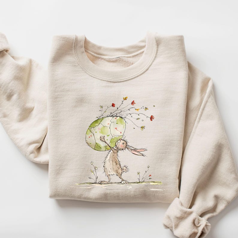 May include: A beige sweatshirt featuring a watercolor illustration of a rabbit carrying a large, cracked egg adorned with flowers. The egg has a green and white design. The rabbit is brown and white, standing on a grassy area.