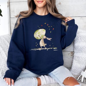 May include: A navy blue long-sleeve sweatshirt featuring a graphic illustration of a brown rabbit standing on its hind legs while holding a large, light green patterned egg above its head, surrounded by small colorful floating flower petals.
