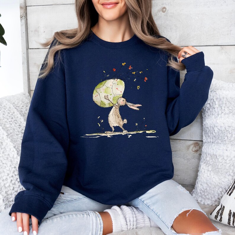 May include: A navy blue long-sleeve sweatshirt featuring a graphic illustration of a brown rabbit standing on its hind legs while holding a large, light green patterned egg above its head, surrounded by small colorful floating flower petals.