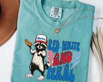 May include: Teal Comfort Colors t-shirt featuring a raccoon wearing a "Merica" hat and holding a hot dog. The shirt has the text "Red White and Feral" with lightning bolt graphics. A gold necklace is draped on the shirt.